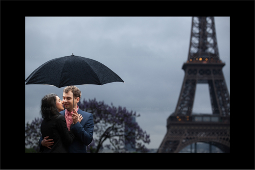 Engaged couple holding tightly and hiding from rain in front of Eiffel Tower